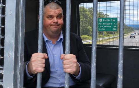 A man in a dark jacket and blue shirt gripping the bars of a cage inside a vehicle, with a Pacific Highway road sign visible through the window behind him.