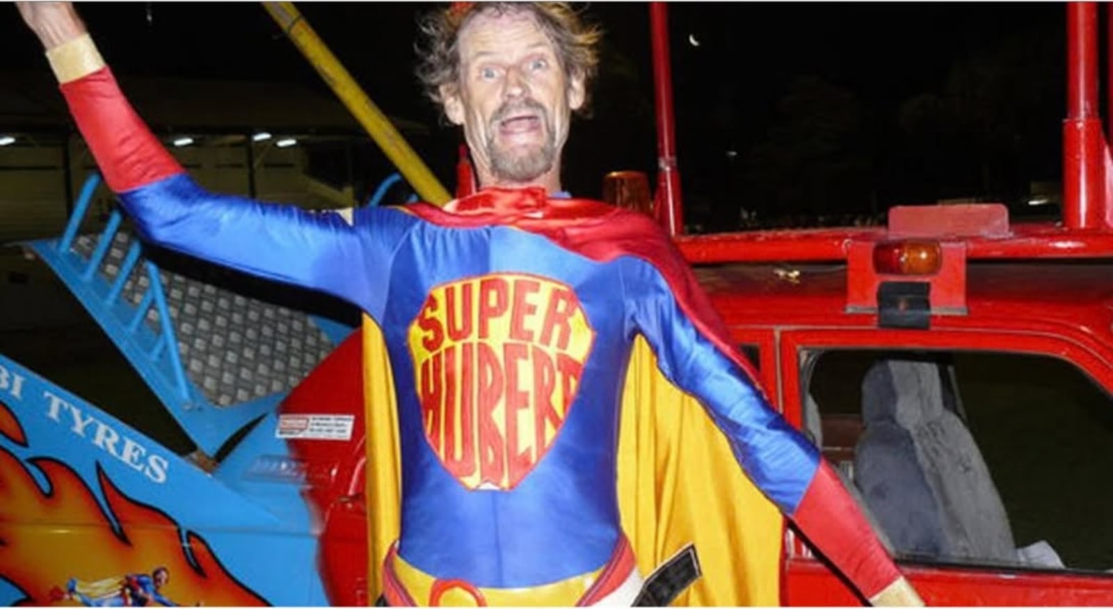 A man in a blue and red superhero costume with the words "Super Hubert" on the chest, standing with his arms out in front of a red truck.