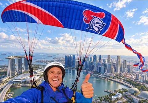 A paraglider under a red and blue Knights canopy giving a thumbs up above the Singapore skyline.
