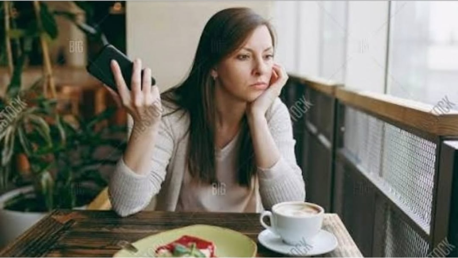 A woman with dark hair sitting at a cafe table by a window, holding a phone to her ear and resting her chin on her hand, with a coffee and a plate of food in front of her.