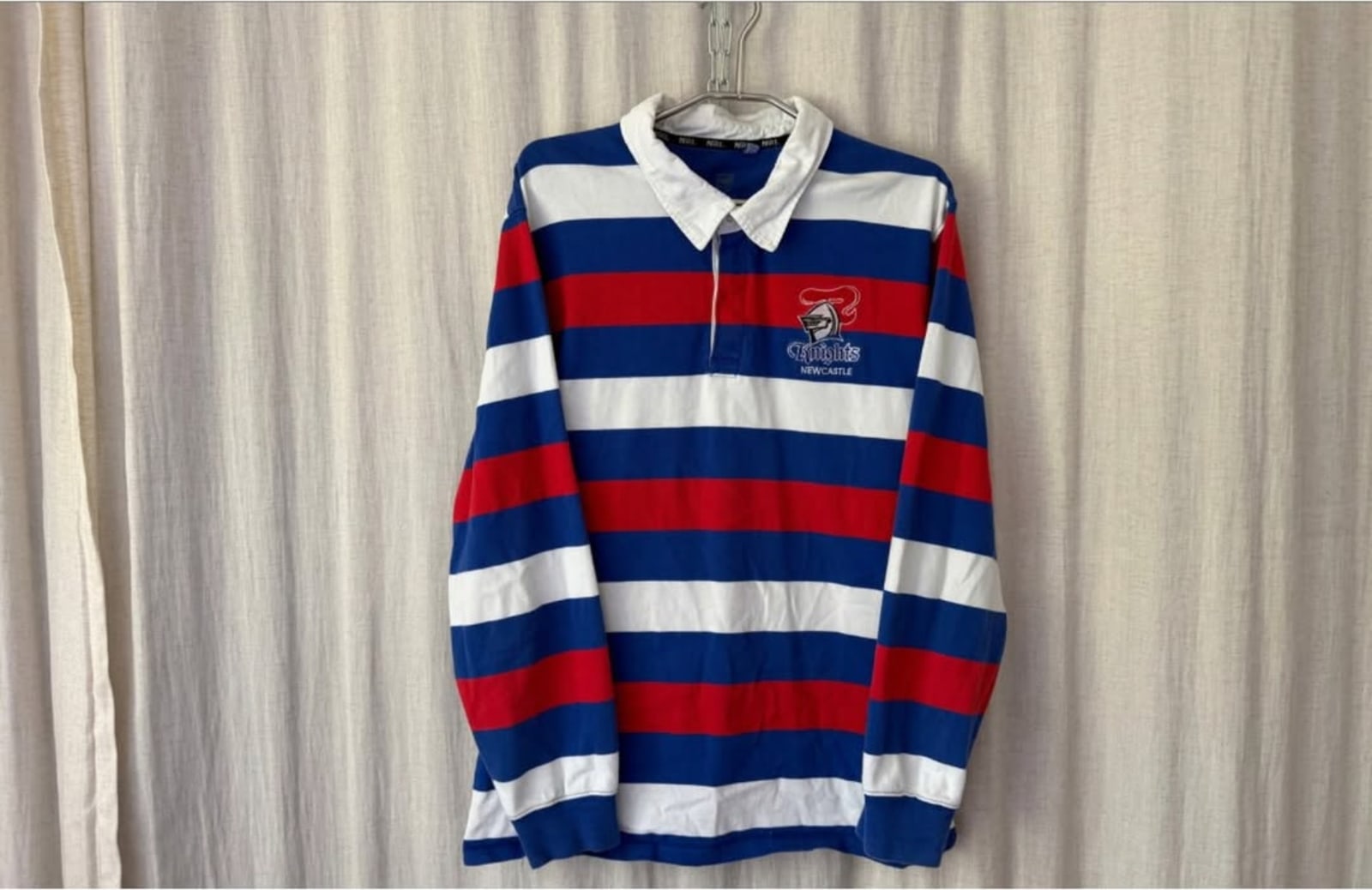 A red, white and blue striped rugby jersey with a Knights badge hanging in front of a cream curtain.