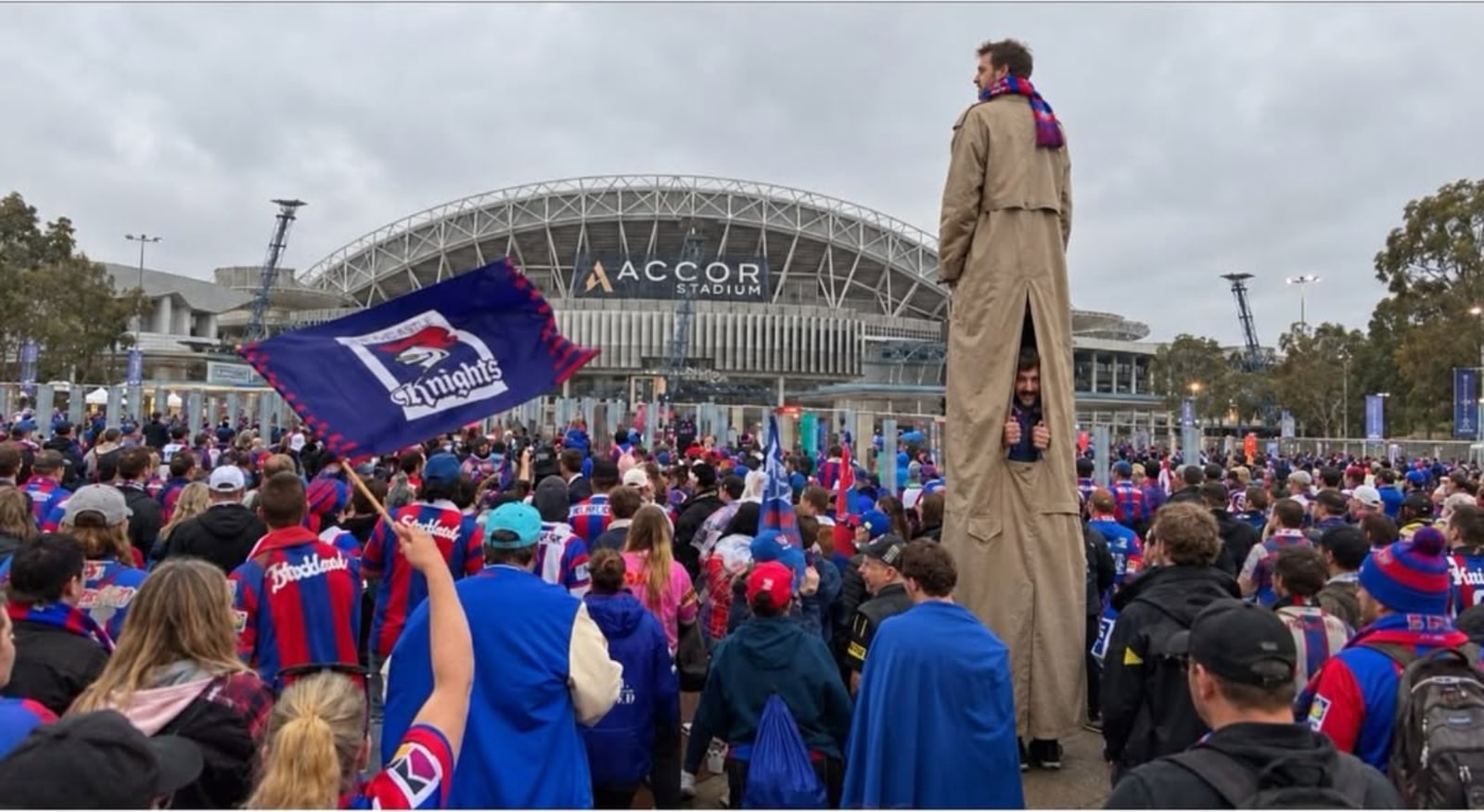 A tall figure on stilts in a long coat standing above a crowd of fans waving Knights flags outside a stadium.