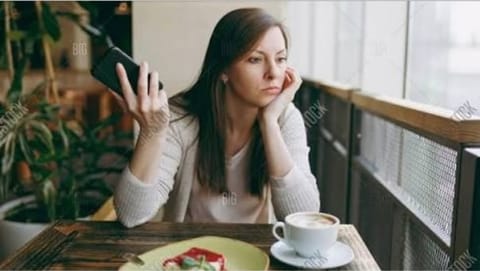 A woman with dark hair sitting at a cafe table by a window, holding a phone to her ear and resting her chin on her hand, with a coffee and a plate of food in front of her.