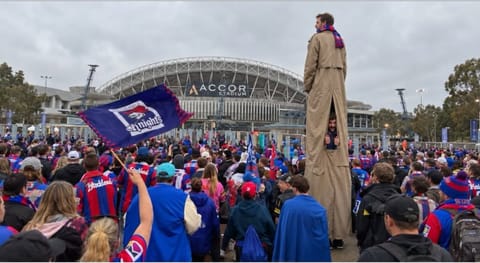 A tall figure on stilts in a long coat standing above a crowd of fans waving Knights flags outside a stadium.