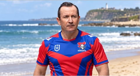 A man in a Knights jersey on the beach with Nobbys headland behind him.
