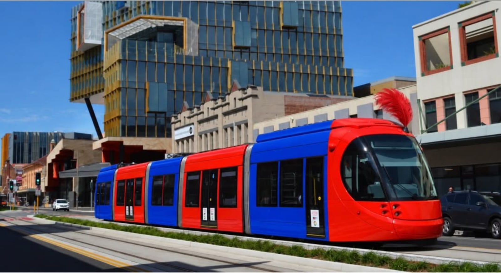 A red and blue light-rail tram running along a city street past a glass office building.