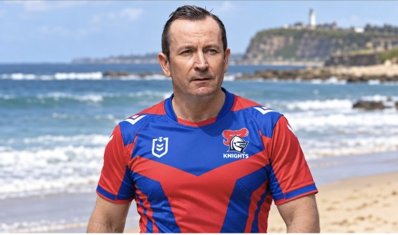 A man in a Knights jersey on the beach with Nobbys headland behind him.